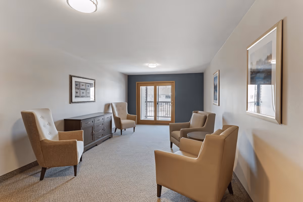 A cozy senior living common area with four beige armchairs arranged around a small round table. The room has light-colored walls with framed artwork, a dark wooden sideboard, carpeted floor, and a set of glass doors leading to an outdoor balcony.