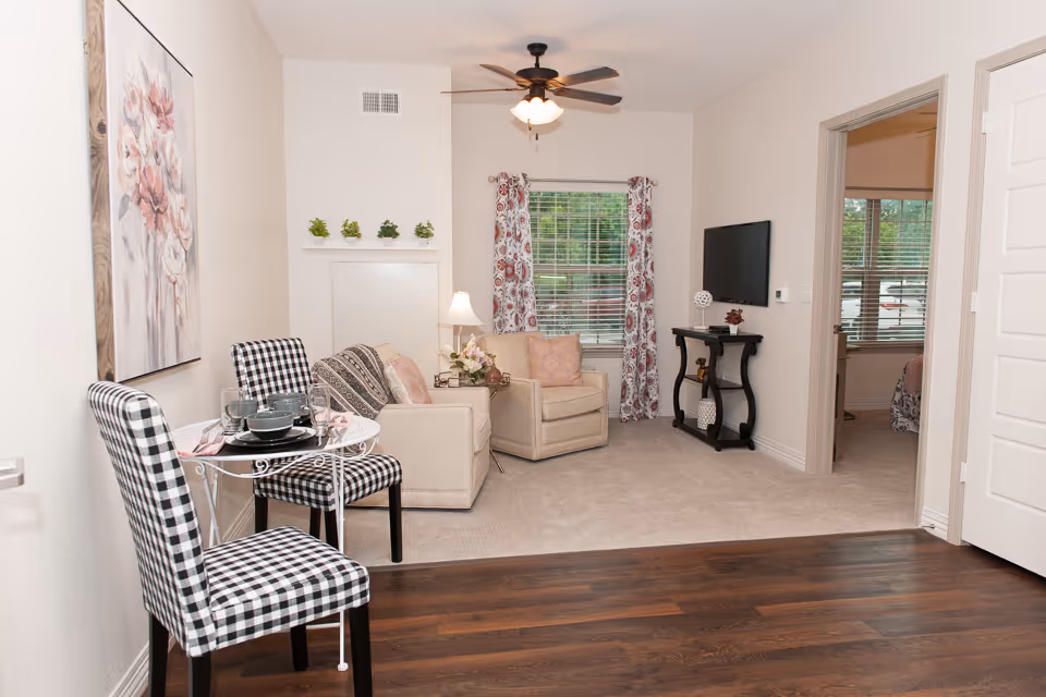 A cozy senior living room with beige walls and carpeted floor. The room features a beige armchair and loveseat with patterned pillows, a small black table with decorative items, a wall-mounted flat-screen TV, and a window with floral curtains. In the foreground, there is a small round dining table set for two with black and white checkered chairs. A ceiling fan with lights hangs above, and a floral painting is on the left wall. A doorway leads to a bedroom with a window and bed visible.