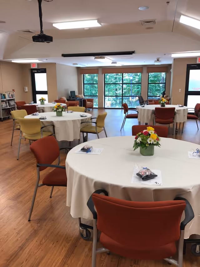 A bright communal dining/activity room with round tables covered in white tablecloths, chairs, floral centerpieces, and large windows showing trees outside.