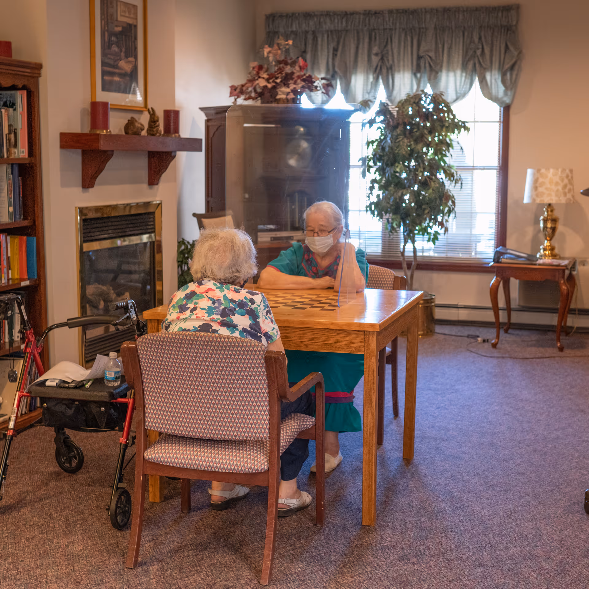Two elderly women sitting across from each other at a wooden table with a clear protective divider between them in a cozy room. One woman is wearing a face mask and a teal dress, while the other has a floral shirt and a walker nearby. The room has a fireplace, bookshelf, potted plant, and a window with curtains letting in natural light.