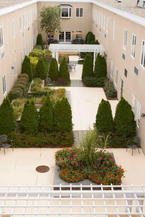An outdoor courtyard between beige buildings with hedges, planters, and seating.