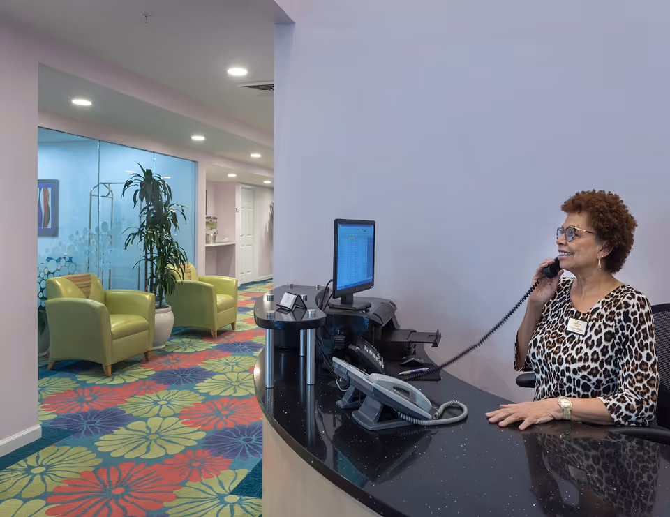 Reception desk in a senior living facility with a staff member answering the phone and a waiting area with green chairs and a colorful floral carpet.