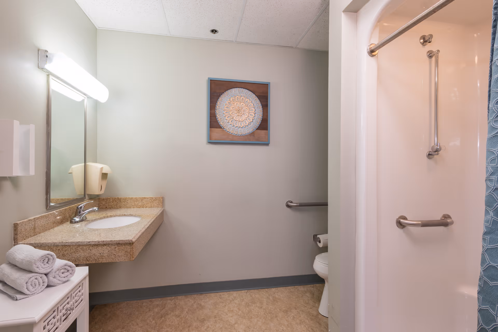 A clean bathroom with a beige countertop and sink, a large mirror with a light above it, a white cabinet with three rolled towels, a toilet with a grab bar, and a shower with grab bars and a blue patterned curtain. A decorative framed artwork hangs on the wall.
