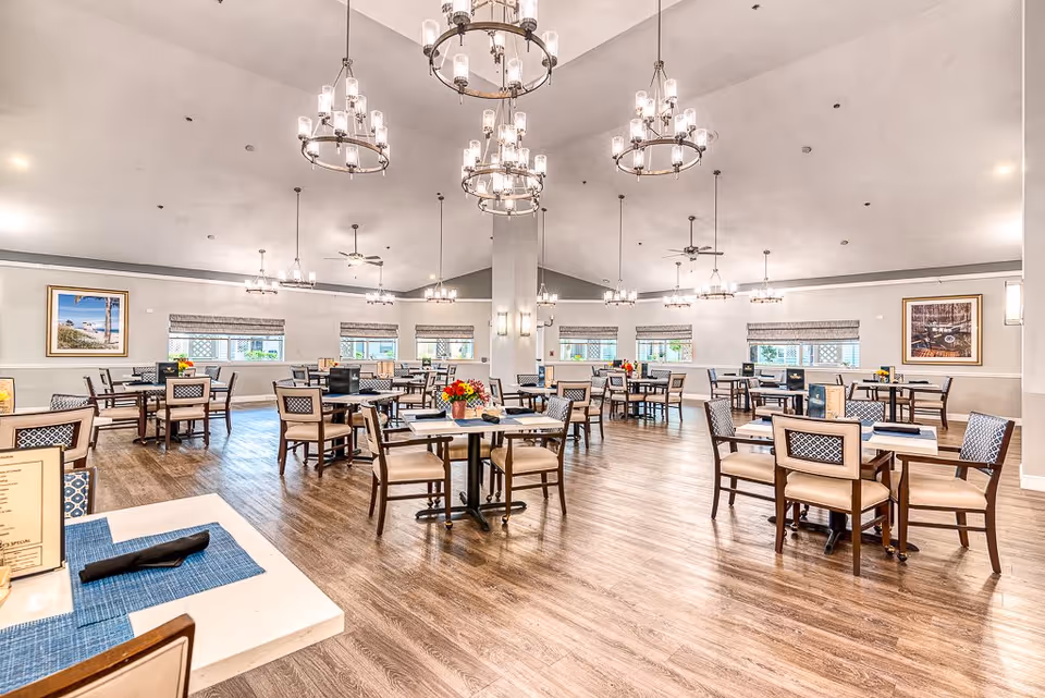 A spacious and well-lit dining room with multiple tables and chairs arranged neatly. The room features wooden flooring, large windows with blinds, and several elegant chandeliers hanging from the ceiling. There are framed pictures on the walls and flower arrangements on some tables.