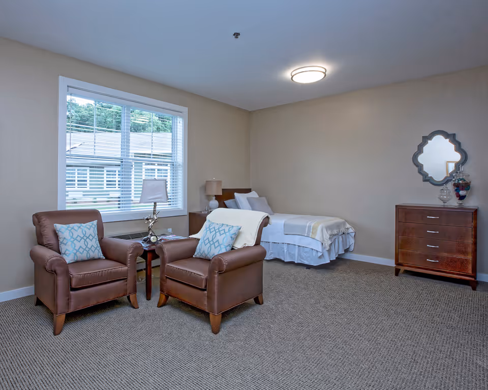 A senior living facility bedroom with a single bed covered in white and beige bedding, two brown armchairs with blue patterned pillows, a small wooden side table with a lamp and clock, a wooden dresser with a decorative mirror above it, and a large window with white blinds showing an exterior view.