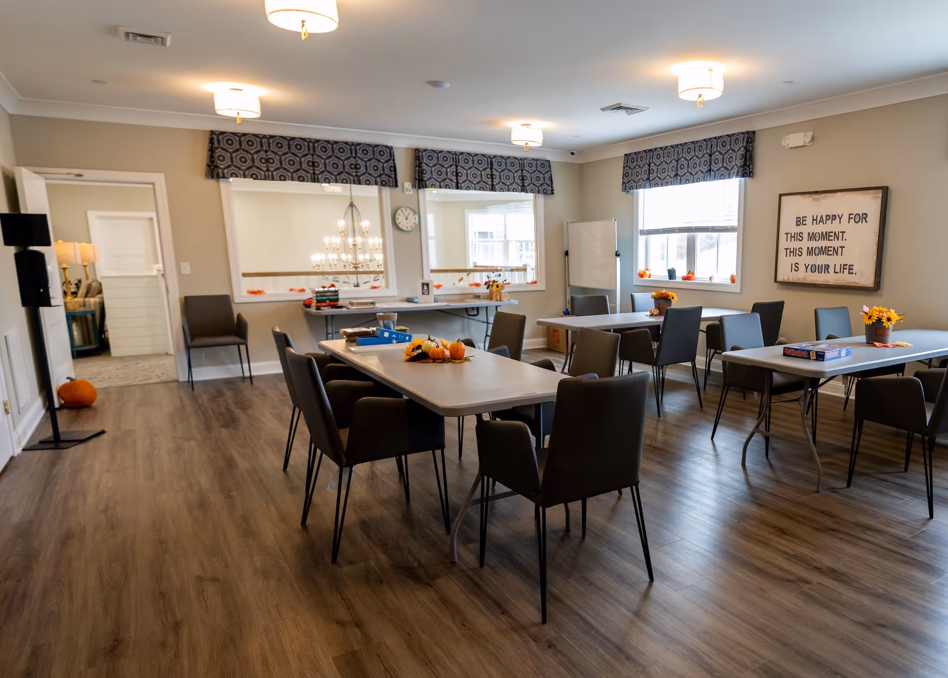 A bright and spacious common room with multiple tables and chairs arranged for group activities. The tables have small decorative pumpkins and flowers. There are large windows with patterned valances letting in natural light. A whiteboard stands near the windows, and a framed sign on the wall reads 'Be happy for this moment. This moment is your life.' The room has wood flooring and ceiling lights.