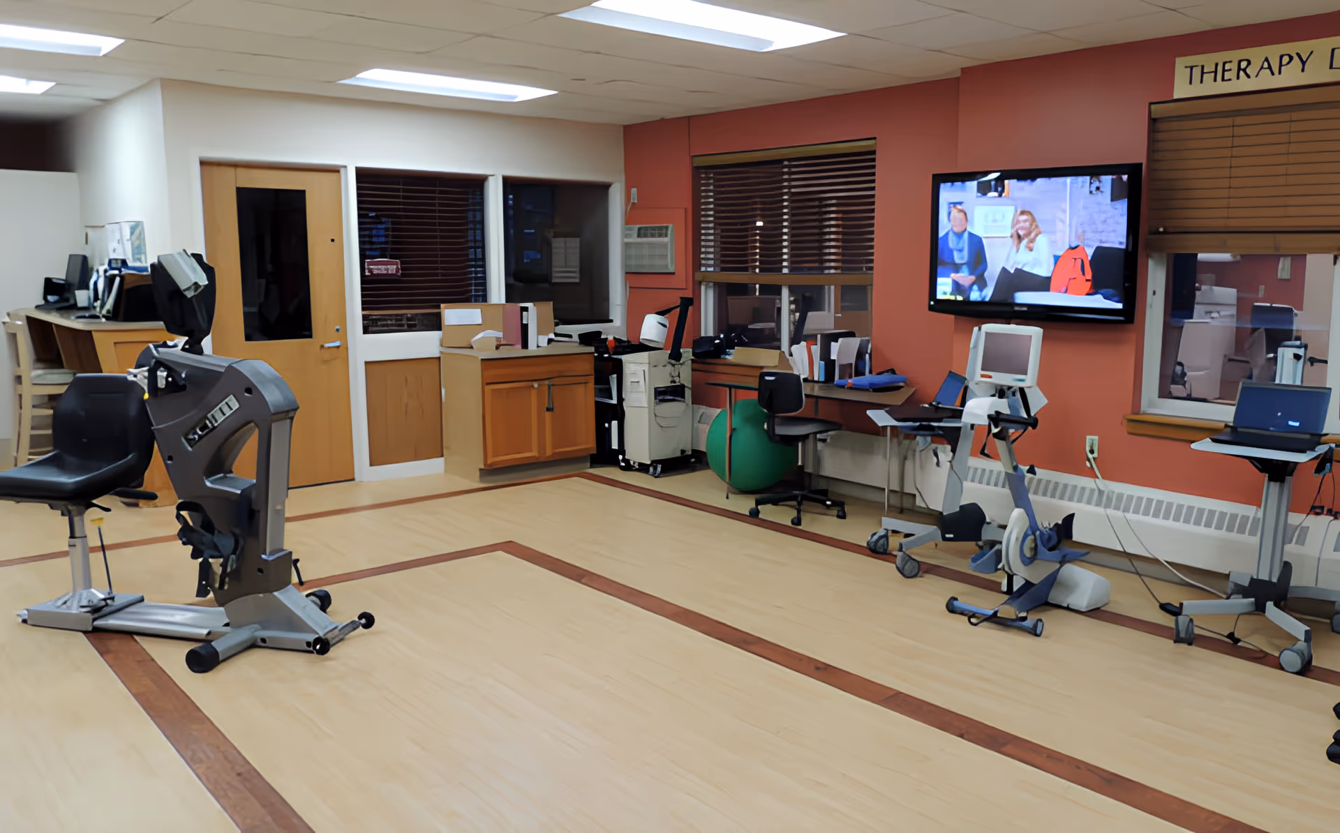 A bright rehabilitation therapy room with exercise machines, desks, a mounted TV, and wood-patterned flooring.
