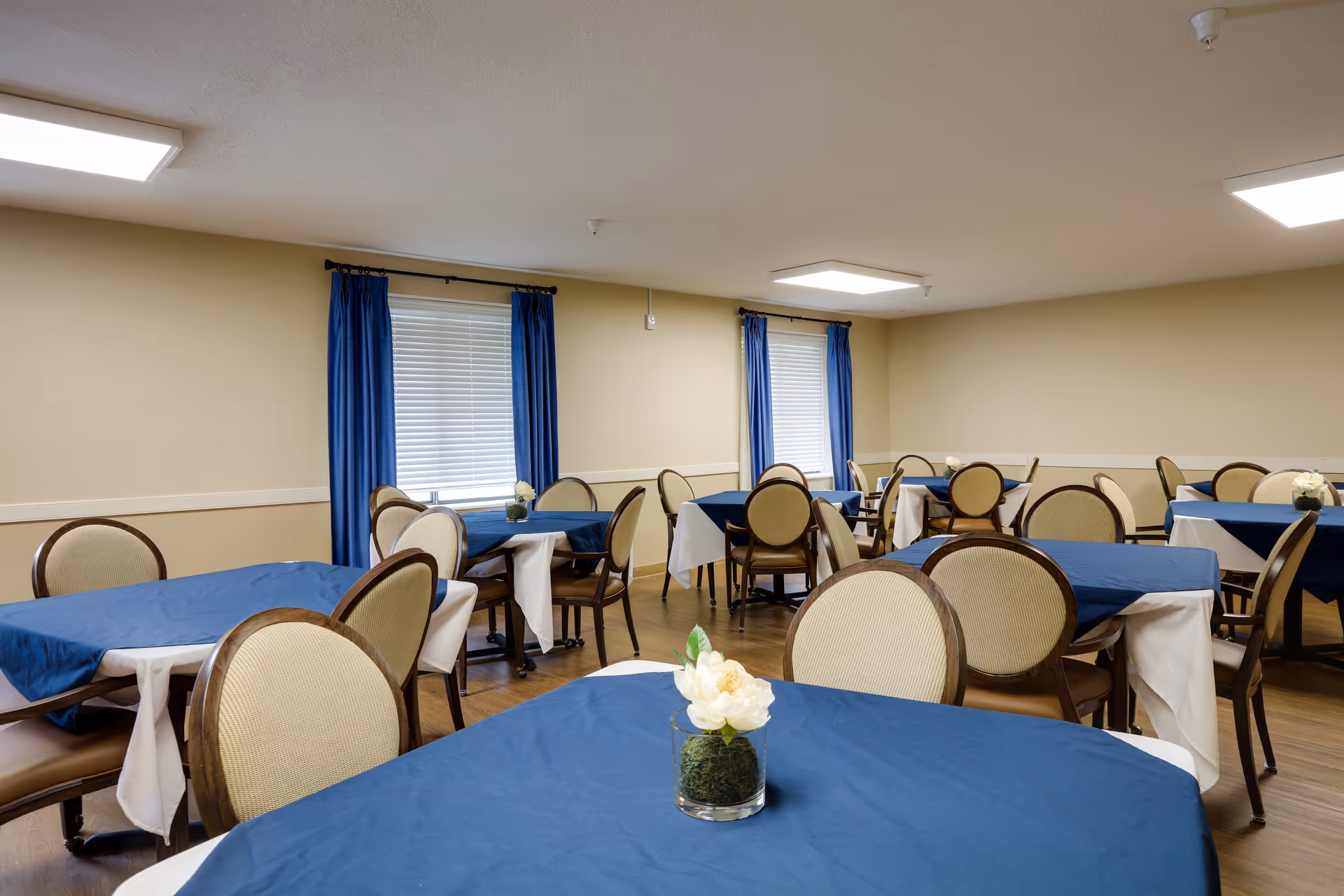 A dining room with multiple tables covered in blue tablecloths and white underlays. Each table is surrounded by beige cushioned chairs with wooden frames. The room has beige walls, two windows with blue curtains, and ceiling lights providing bright illumination. Small flower arrangements are placed on the tables.