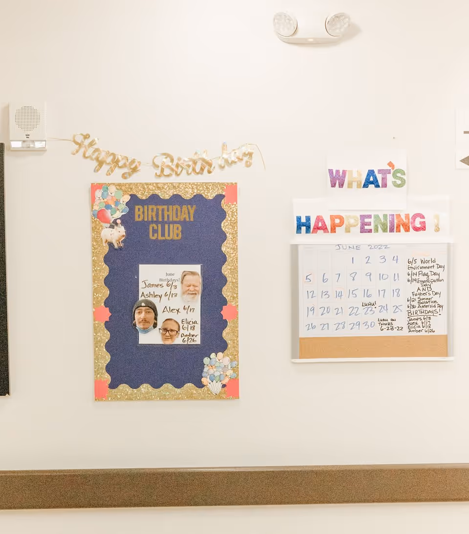A bulletin board display on a wall featuring a 'Happy Birthday' banner, a 'Birthday Club' board with photos and names of residents with June birthdays, and a 'What's Happening!' calendar for June 2022 listing events and birthdays.