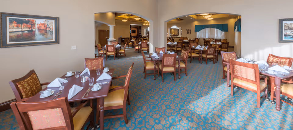 A spacious dining room with multiple wooden tables and chairs arranged neatly. Each table is set with white napkins, cups, and glasses. The room has patterned blue and beige carpeting, beige walls, and framed artwork hanging on the walls. Large archways lead to another section of the dining area with more tables and chairs.