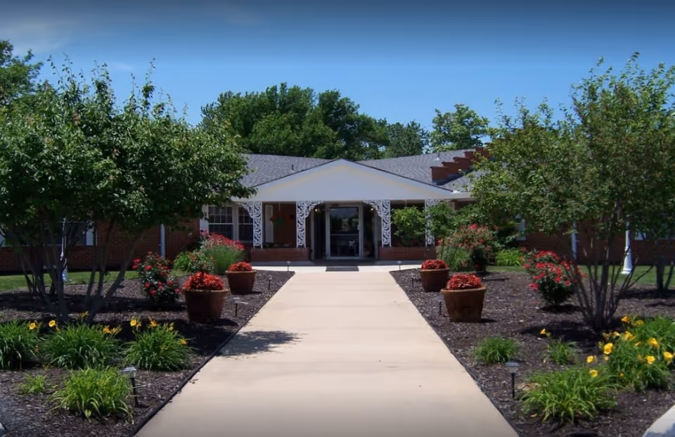Front exterior view of Heritage Avonlea of Olathe facility with a paved walkway leading to the entrance, surrounded by landscaped garden beds with trees, shrubs, and potted red flowers under a clear blue sky.