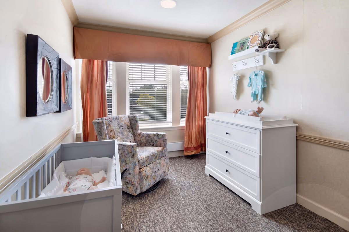 A small nursery room with a gray crib containing a baby doll, a patterned armchair near a window with red curtains, and a white dresser with a changing pad on top and another baby doll lying on it. Above the dresser is a white shelf with hooks holding baby clothes and some decorative items.