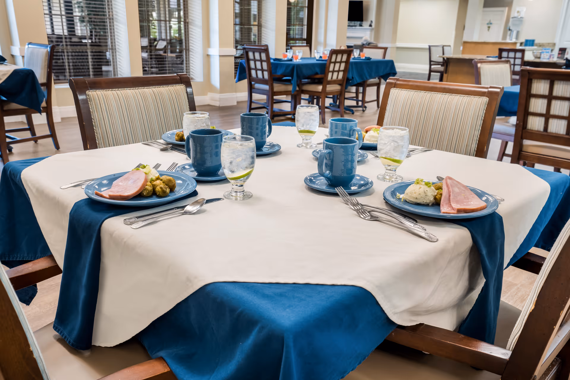 A dining table set for four with blue plates containing ham, mashed potatoes, and Brussels sprouts. Each place setting has a blue coffee cup, a glass of water with a lemon slice, and silverware. The table is covered with a white tablecloth over a blue one. The background shows more tables and chairs in a well-lit dining room with large windows.