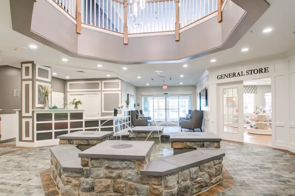 Interior view of a senior living facility lobby with a stone water fountain in the center, a reception desk to the left, seating area with chairs in the background, and a doorway labeled 'GENERAL STORE' on the right.