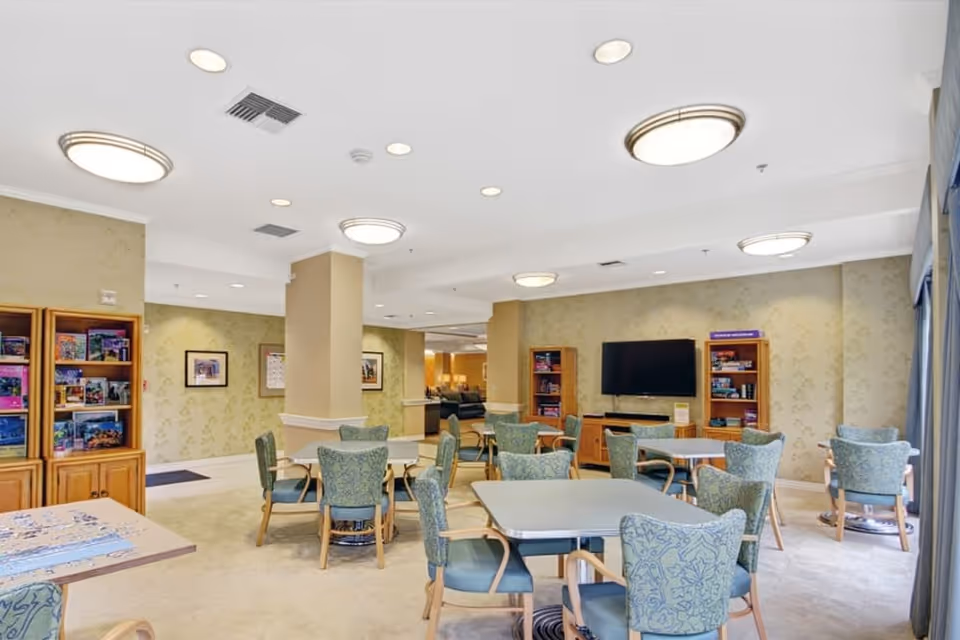 A well-lit common area in a senior living facility featuring multiple tables with green patterned chairs arranged around them. The room has beige walls with subtle floral patterns, ceiling lights, and a flat-screen TV mounted on the wall flanked by wooden bookshelves filled with board games and books. Large windows with curtains allow natural light to enter the space.