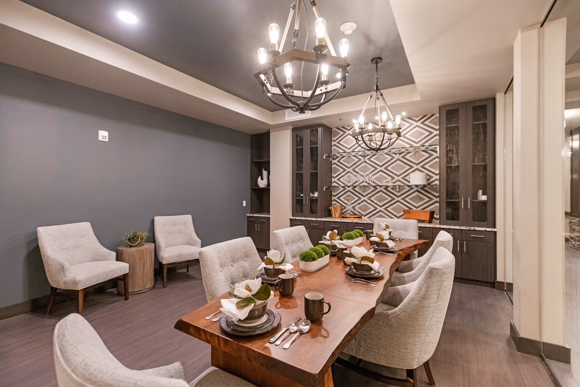 A modern dining room with a long wooden table set for six people. The table is decorated with white flowers and green plants. There are upholstered chairs around the table and two chandeliers hanging from the ceiling. The back wall features a geometric patterned backsplash with glass shelves and dark cabinetry.