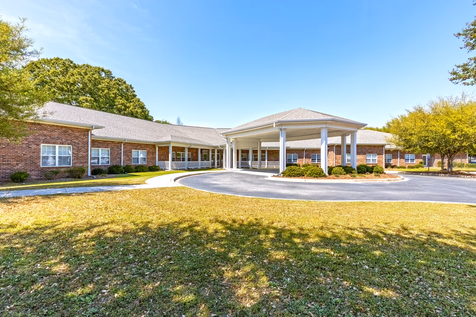 Exterior view of a single-story brick building with a covered entrance driveway, surrounded by green grass and trees under a clear blue sky.
