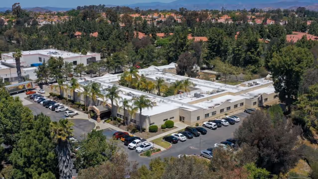 Aerial view of Carmel Mountain Rehabilitation And Healthcare Center, a single-story building with a white roof surrounded by trees and parking lots filled with cars. The facility is located in a suburban area with hills and residential houses in the background.