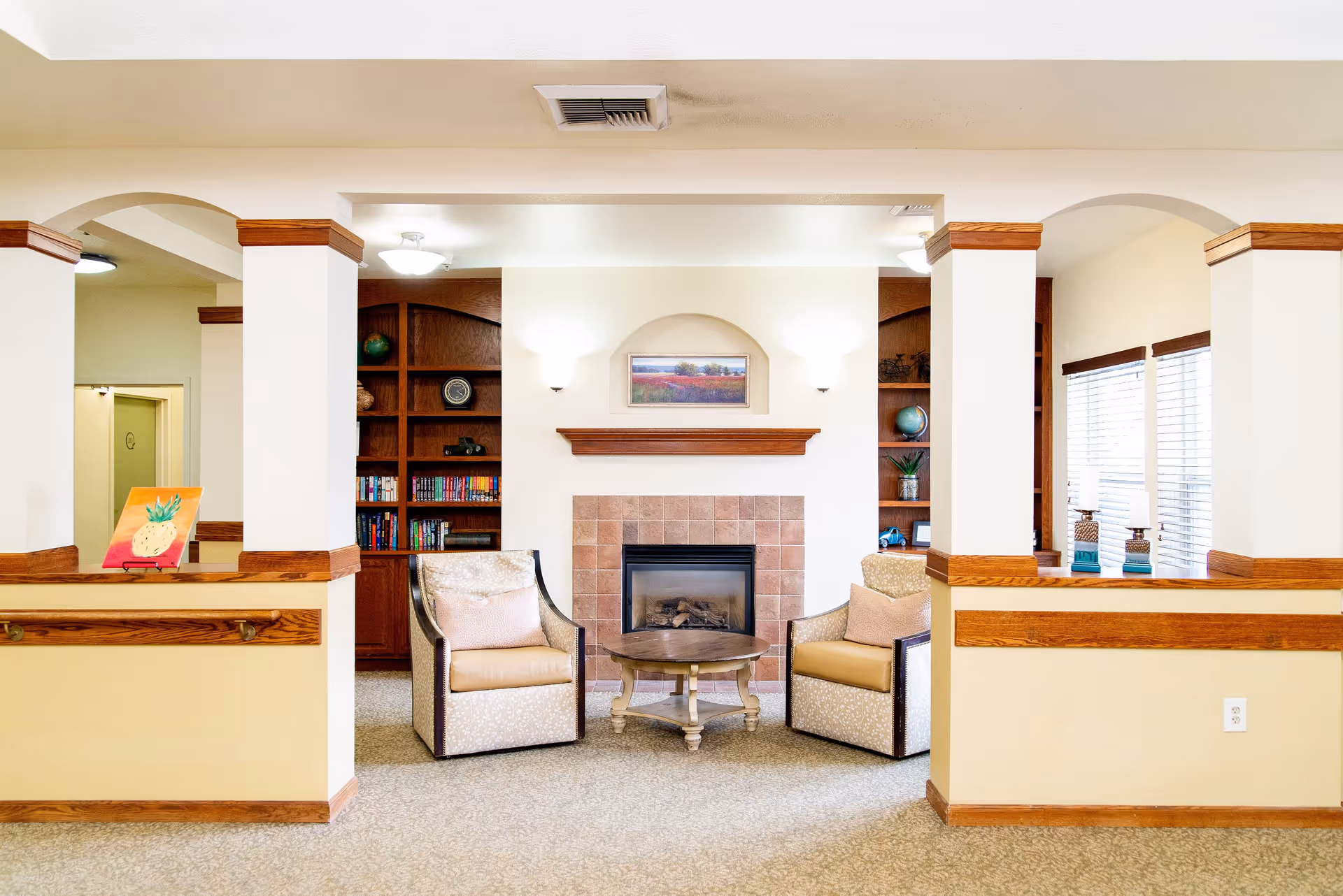 A cozy sitting area in a senior living facility featuring two cushioned armchairs with pillows, a round wooden coffee table, and a fireplace with a tiled surround. Built-in wooden bookshelves flank the fireplace, holding books, globes, and decorative items. The room has beige walls with wooden trim and carpeted flooring, with windows on the right side letting in natural light.