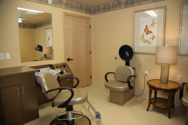 Interior of a hair salon area in a senior living facility featuring two salon chairs, a hair washing station, a large mirror, a wooden door, a side table with a lamp, and a framed butterfly artwork on the wall.