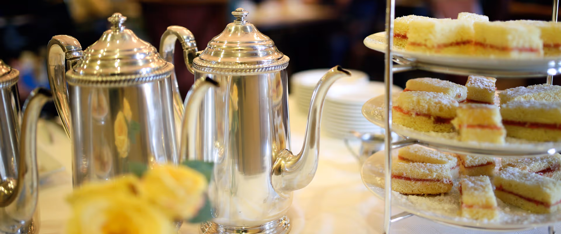 A close-up view of a tea service setup featuring three polished silver teapots and a three-tiered tray filled with small square sponge cakes dusted with powdered sugar and layered with jam, set on a white tablecloth with a blurred yellow flower in the foreground.