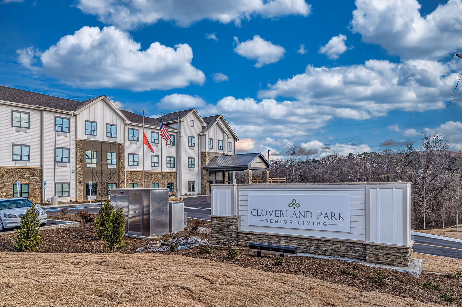 Exterior view of Cloverland Park Senior Living facility showing a three-story building with stone and white siding, a parking lot with cars, two flagpoles with flags, and a large sign with the facility name in front. The sky is partly cloudy.