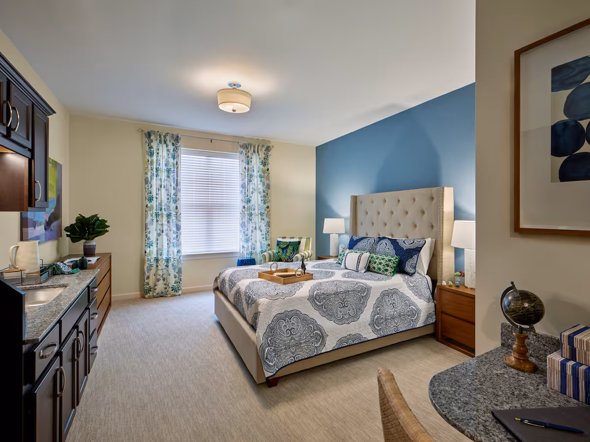 Well-lit furnished bedroom with a tufted headboard bed, blue accent wall, floral curtains, bedside tables, and a small kitchenette area.