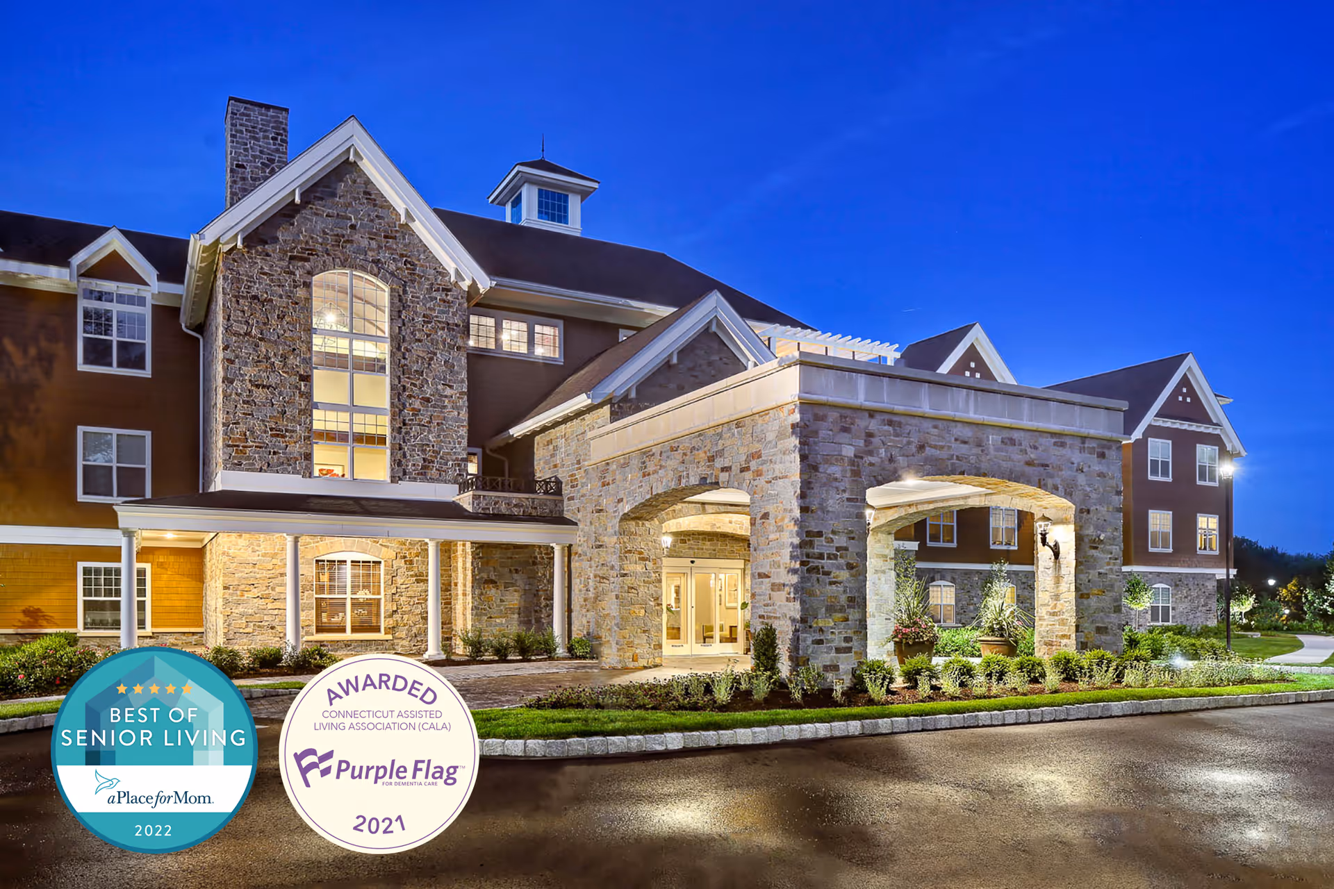 Front exterior of a well-lit stone-and-brick senior living facility at dusk with an arched entrance and award badges in the lower-left corner.