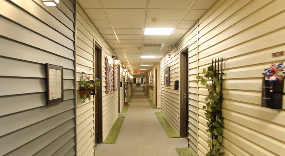 Long indoor corridor lined with home-style siding, apartment doors, wall planters and overhead fluorescent lights.