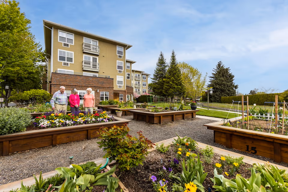 Three elderly people standing and enjoying a garden with raised flower beds filled with colorful flowers and plants. Behind them is a multi-story residential building with beige siding and brick accents. The sky is partly cloudy and there are trees and greenery surrounding the garden area.