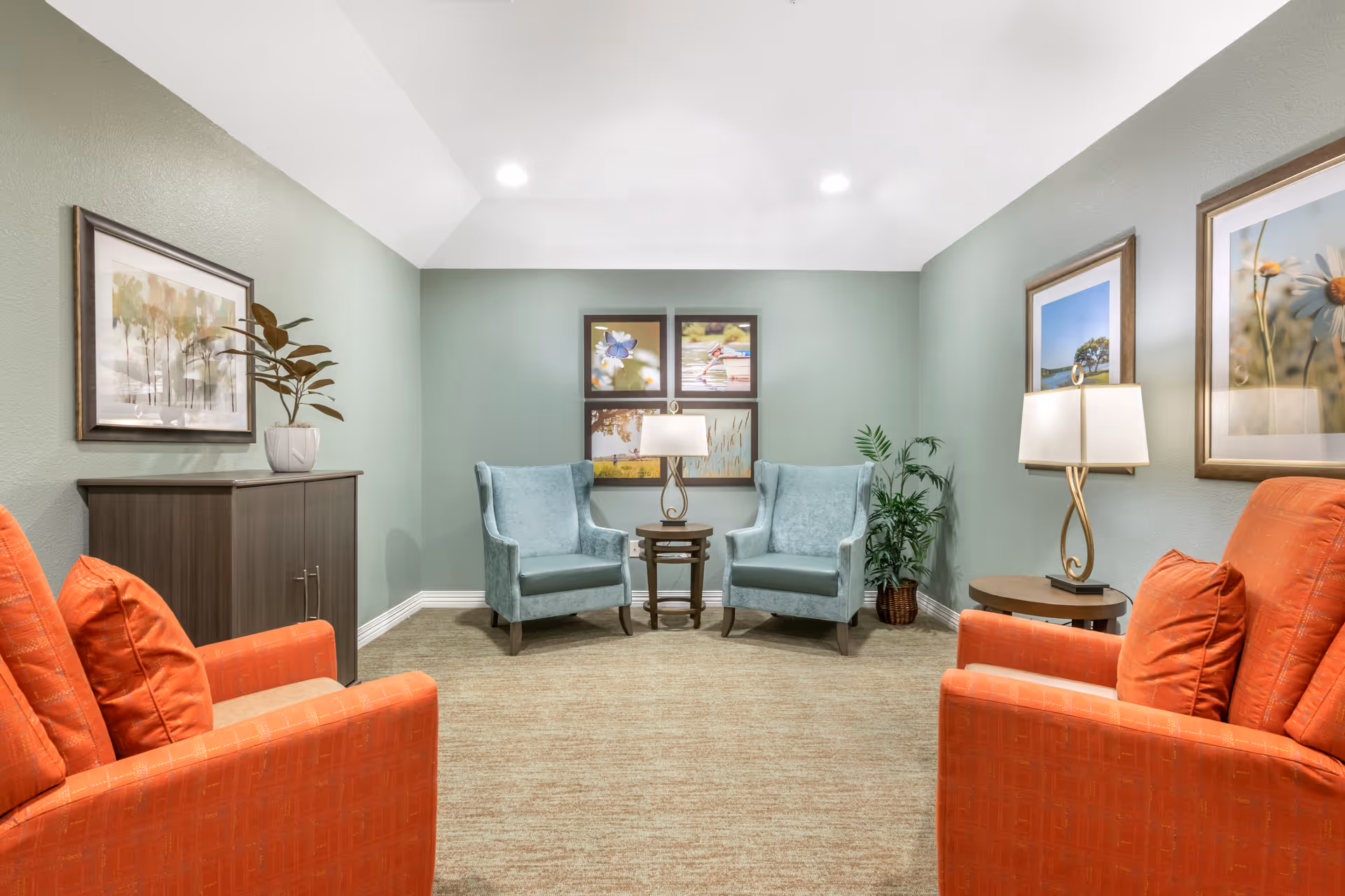 A cozy sitting area with two light blue armchairs facing two orange armchairs. A small round wooden table with a lamp is placed between the blue chairs. The walls are painted light green and decorated with framed pictures of nature scenes. There is a small cabinet with a potted plant on top on the left side and another potted plant on the right side near the wall.