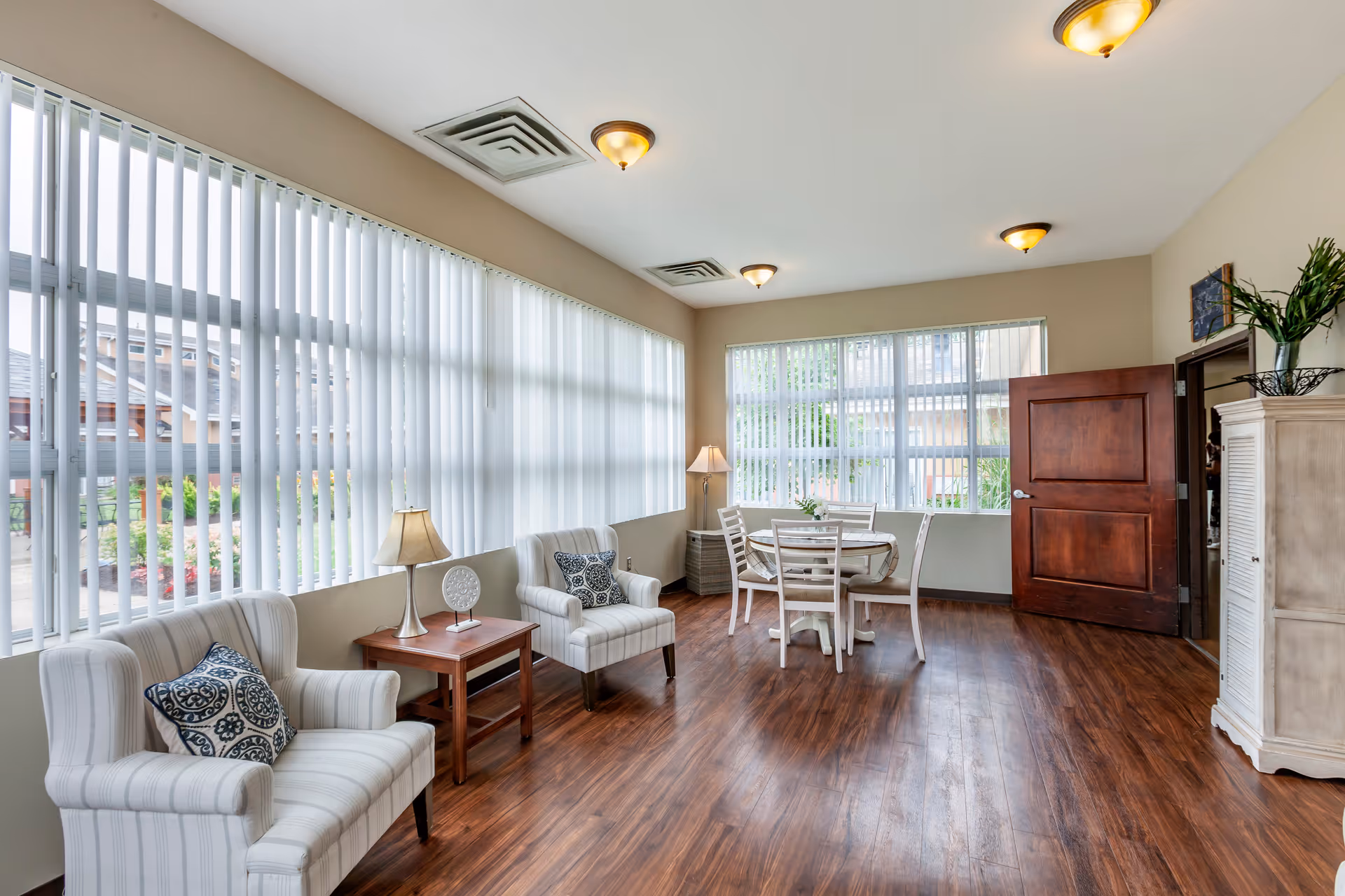 A bright and spacious room with large windows covered by vertical blinds, featuring two upholstered armchairs with patterned cushions, a wooden side table with a lamp and decorative plate, a round dining table with four chairs, wooden flooring, beige walls, and ceiling lights. A wooden door is open, and a white cabinet with a plant on top is visible.