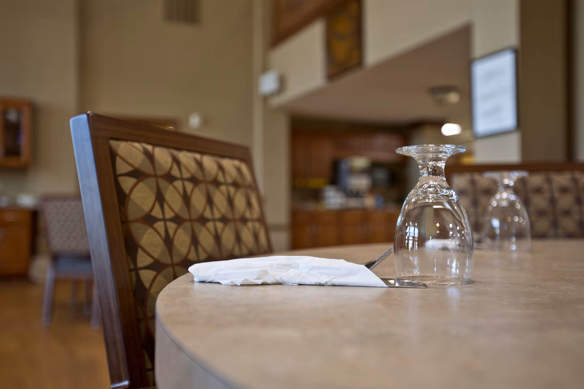Close-up of a dining table in a senior living facility set with a folded napkin, utensils and upside-down wine glasses, with patterned chairs and a blurred dining area in the background.
