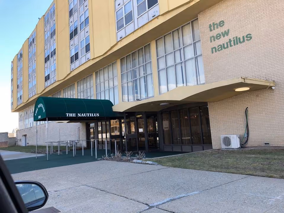 Exterior view of a multi-story building with a green awning labeled 'THE NAUTILUS' over the entrance. The building has large windows and beige brick walls with the words 'the new nautilus' painted on the wall near the entrance. There is a sidewalk and some grass in front of the building.