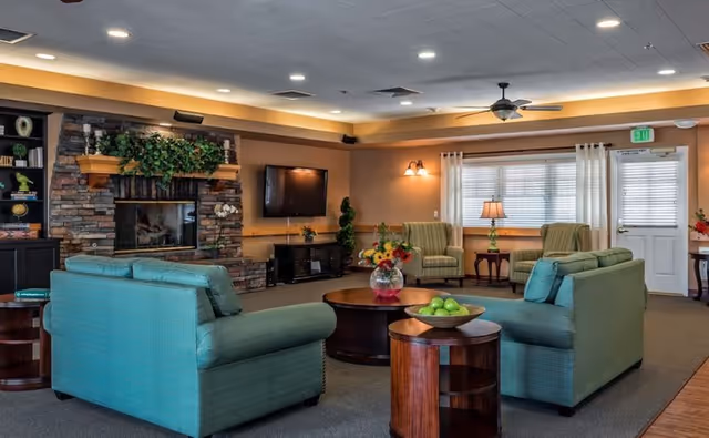 A cozy living room area in a senior living facility with two green upholstered sofas facing each other around a round wooden coffee table. The room features a stone fireplace with greenery on the mantle, a wall-mounted flat-screen TV, and two green armchairs near a window with white curtains. There are wooden side tables with a bowl of green apples and a vase of flowers, ceiling fans, and warm lighting.