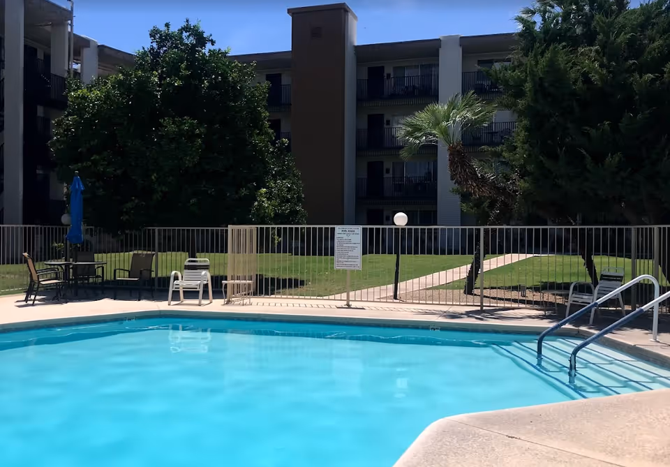 Outdoor swimming pool with clear blue water surrounded by a safety fence. There are several chairs and a table with a closed blue umbrella near the pool. Behind the pool area, there is a grassy lawn with trees and a multi-story residential building in the background.