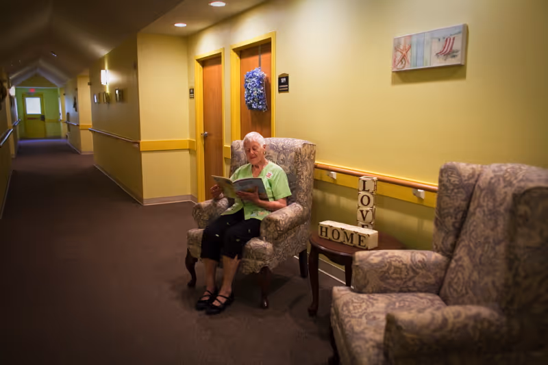 An elderly woman sitting in a patterned armchair in a well-lit hallway of an assisted living facility, reading a book. The hallway has yellow walls with a chair rail, several closed doors, and another armchair nearby. A small table between the chairs holds decorative blocks spelling 'LOVE' and 'HOME'.