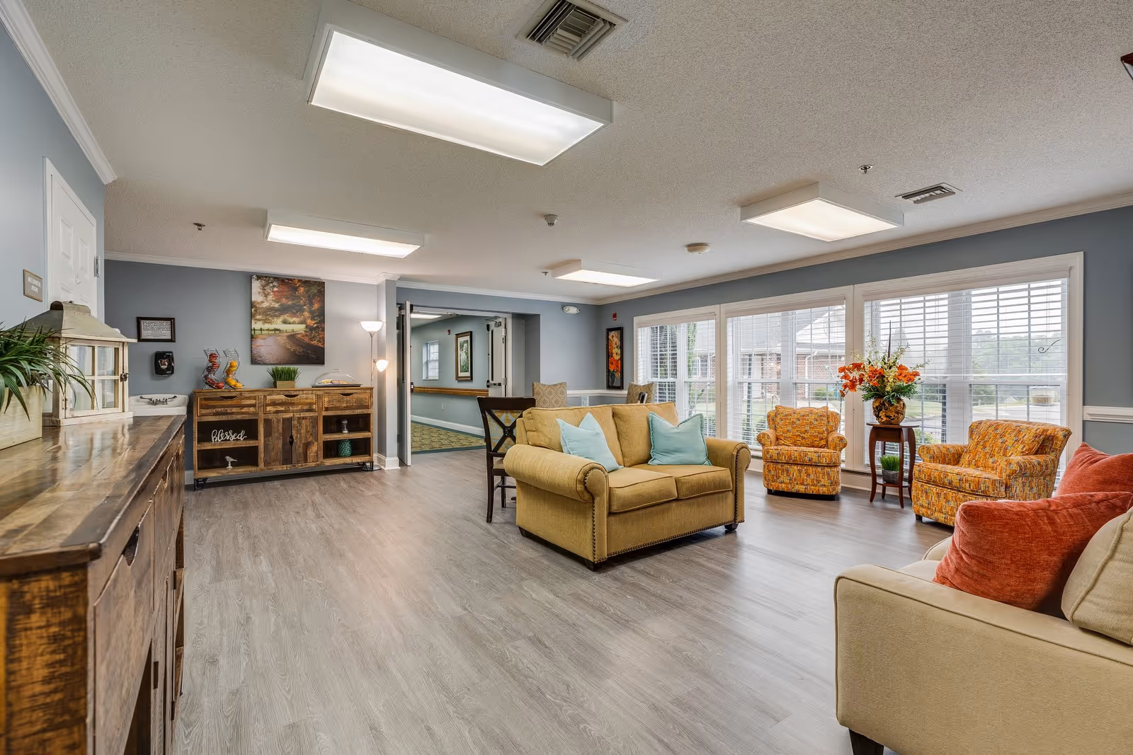 A bright and spacious living room area in an assisted living facility with large windows letting in natural light. The room features a beige loveseat with blue pillows, two patterned armchairs, a beige sofa with red pillows, wooden furniture including a sideboard and a cabinet, and decorative plants and artwork on the walls.