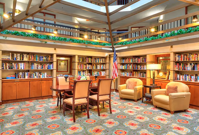 A spacious, well-lit library room with wooden bookshelves filled with books lining the walls. In the center, there is a wooden table with four chairs around it. To the right, there are two cushioned armchairs with a small side table and a lamp between them. An American flag stands near the bookshelves. The room has a patterned carpet and an open ceiling with visible beams and upper-level railings decorated with green plants.