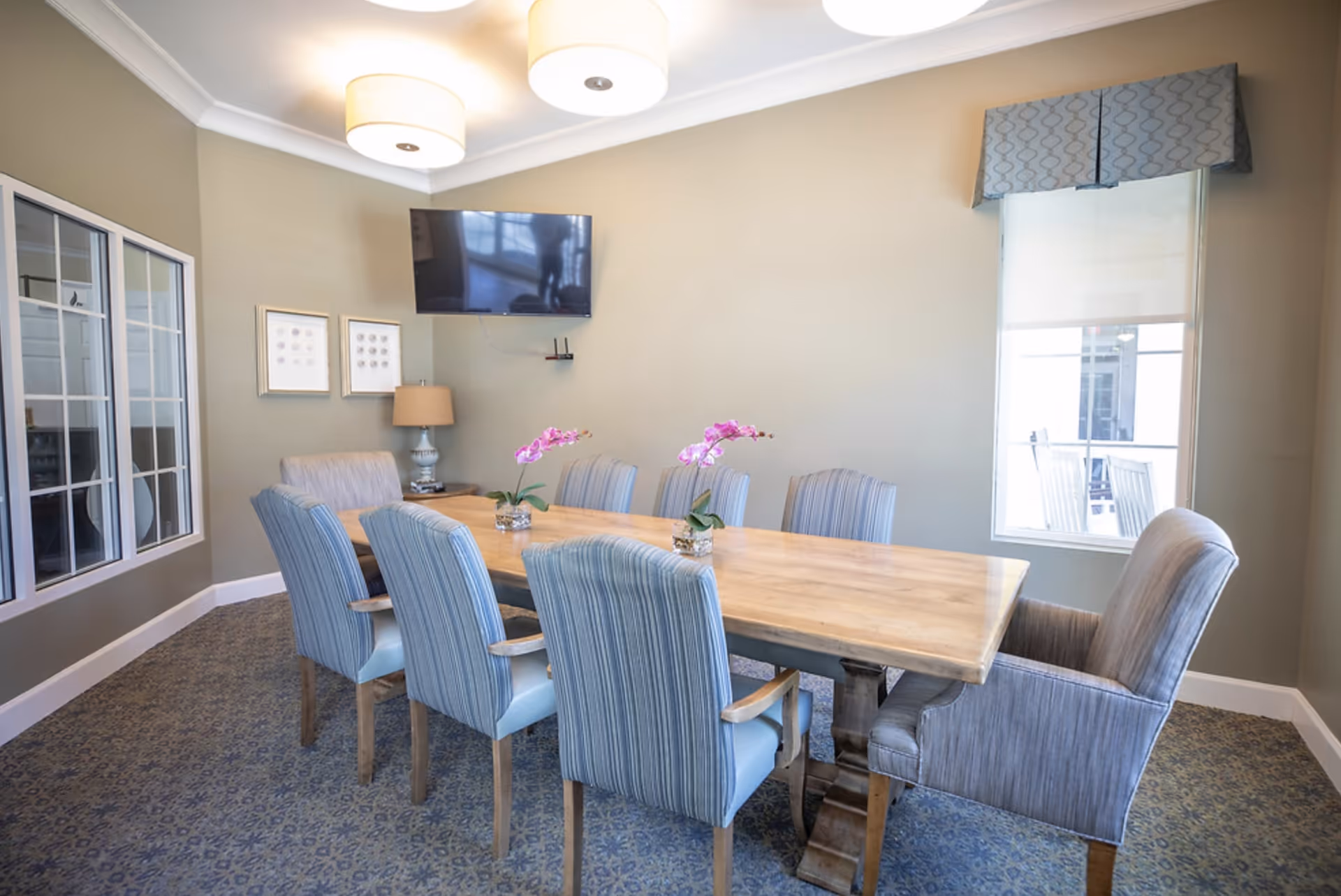A dining room with a long wooden table surrounded by eight upholstered chairs in blue and gray tones. Two small flower arrangements with pink orchids are placed on the table. The room has beige walls, a window with a patterned valance, a wall-mounted flat-screen TV, two framed pictures, a table lamp, and two ceiling lights.