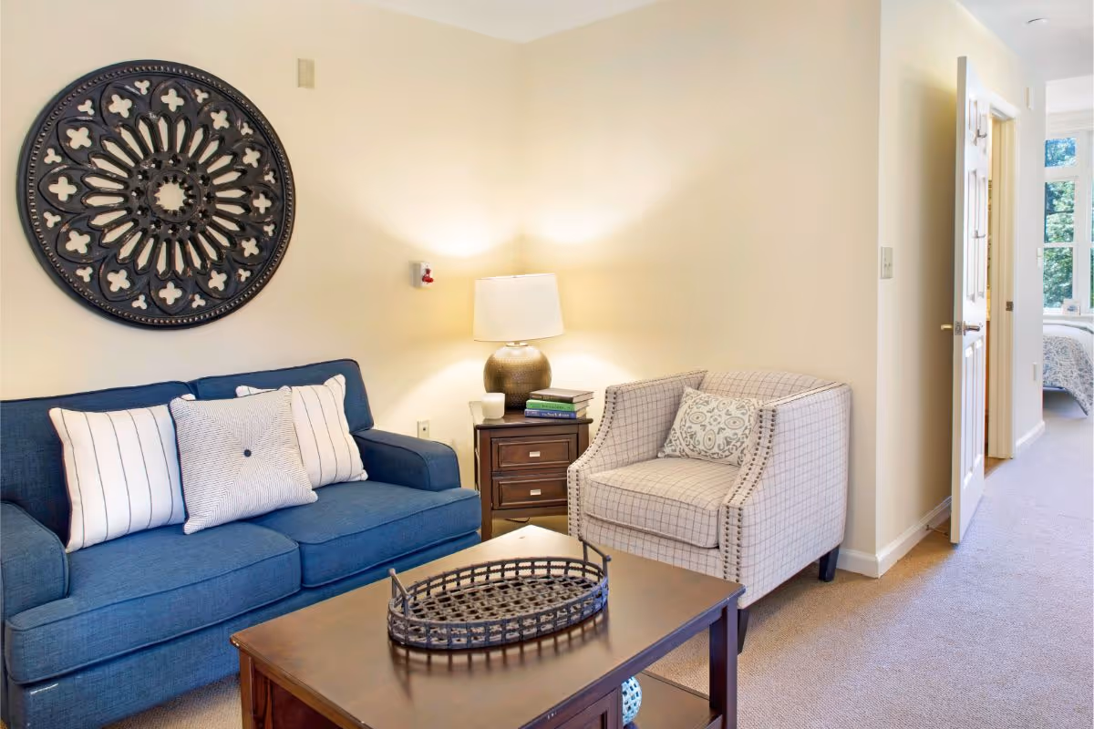 A cozy living room with a blue sofa, patterned armchair, coffee table, table lamp, and decorative wall medallion, with an open doorway leading to a bedroom.