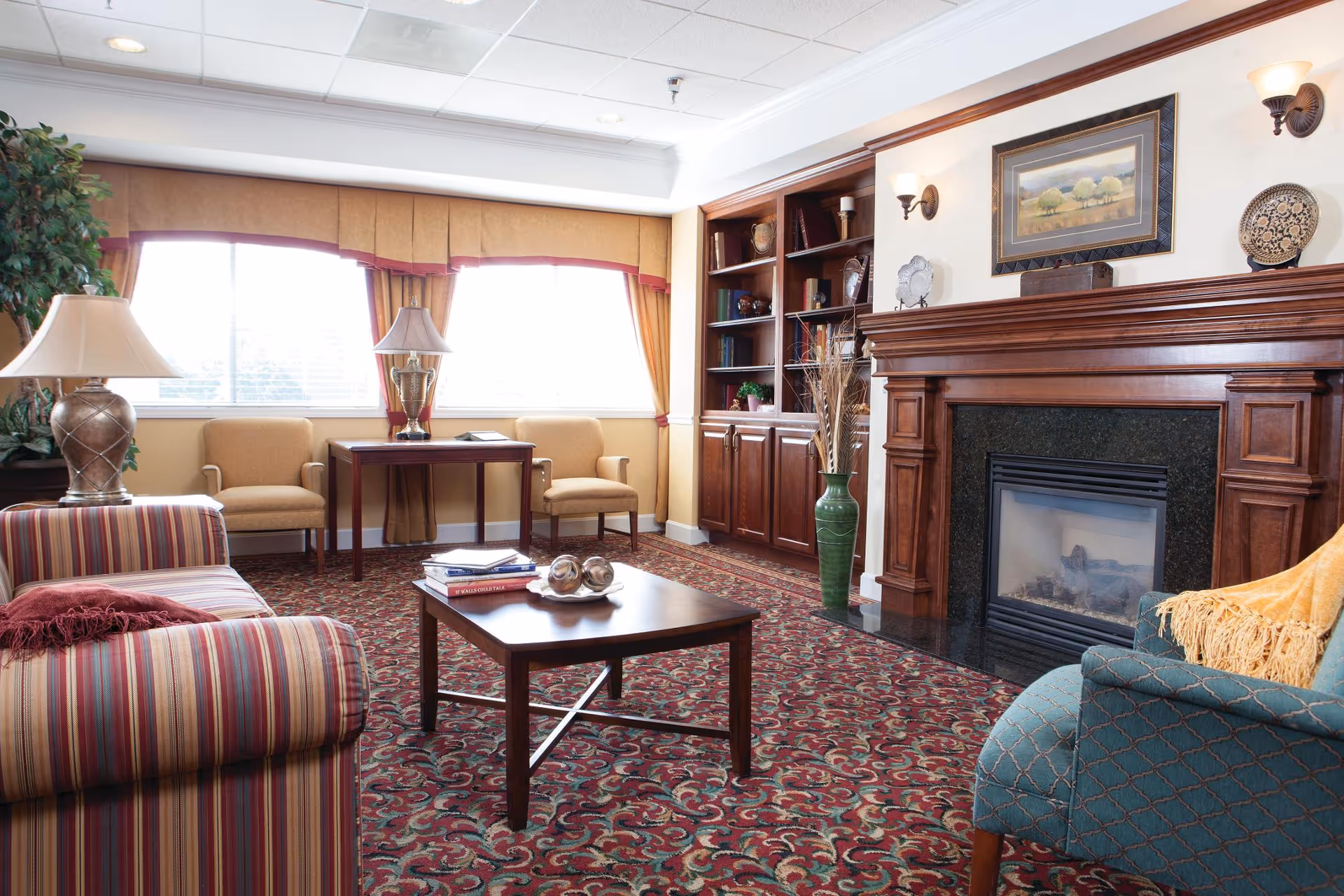 A cozy living room with a striped sofa, a blue armchair with a yellow throw, a wooden coffee table with books and decorative items, two beige chairs by a window with yellow curtains, a wooden bookshelf, and a fireplace with a framed painting above it.