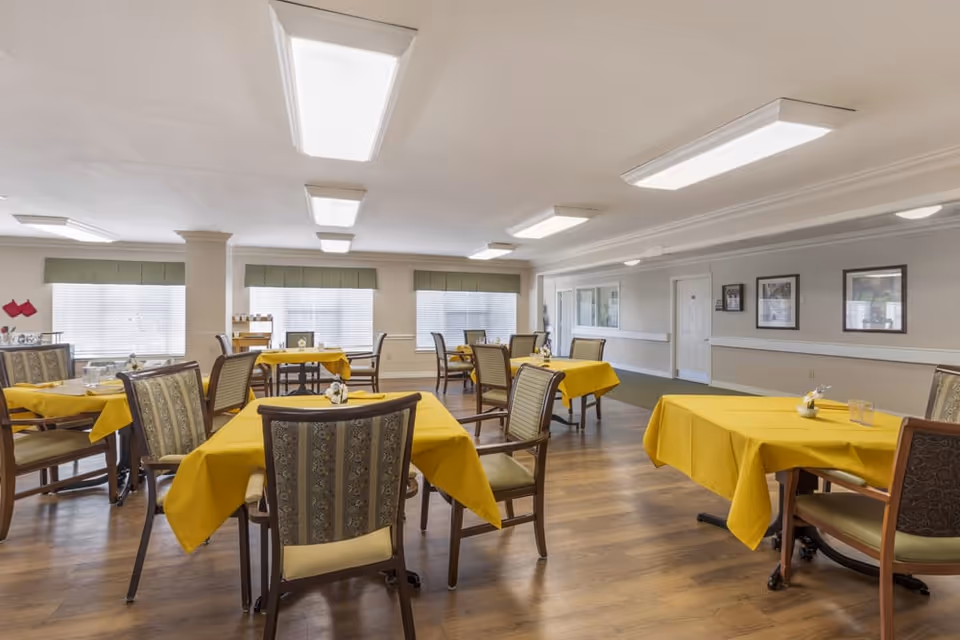 A dining room with multiple tables covered in yellow tablecloths, each surrounded by chairs with patterned upholstery. The room has large windows with blinds, wood flooring, and bright overhead lighting. The walls are decorated with framed pictures.