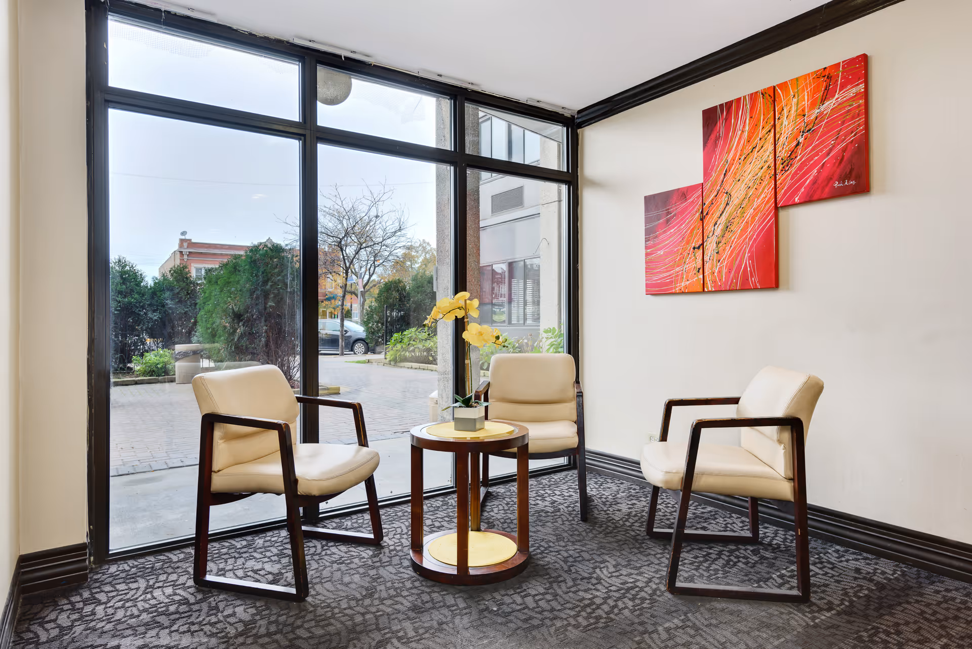 Three beige armchairs arranged around a small round table in a bright lobby seating area with large floor-to-ceiling windows and colorful wall art.