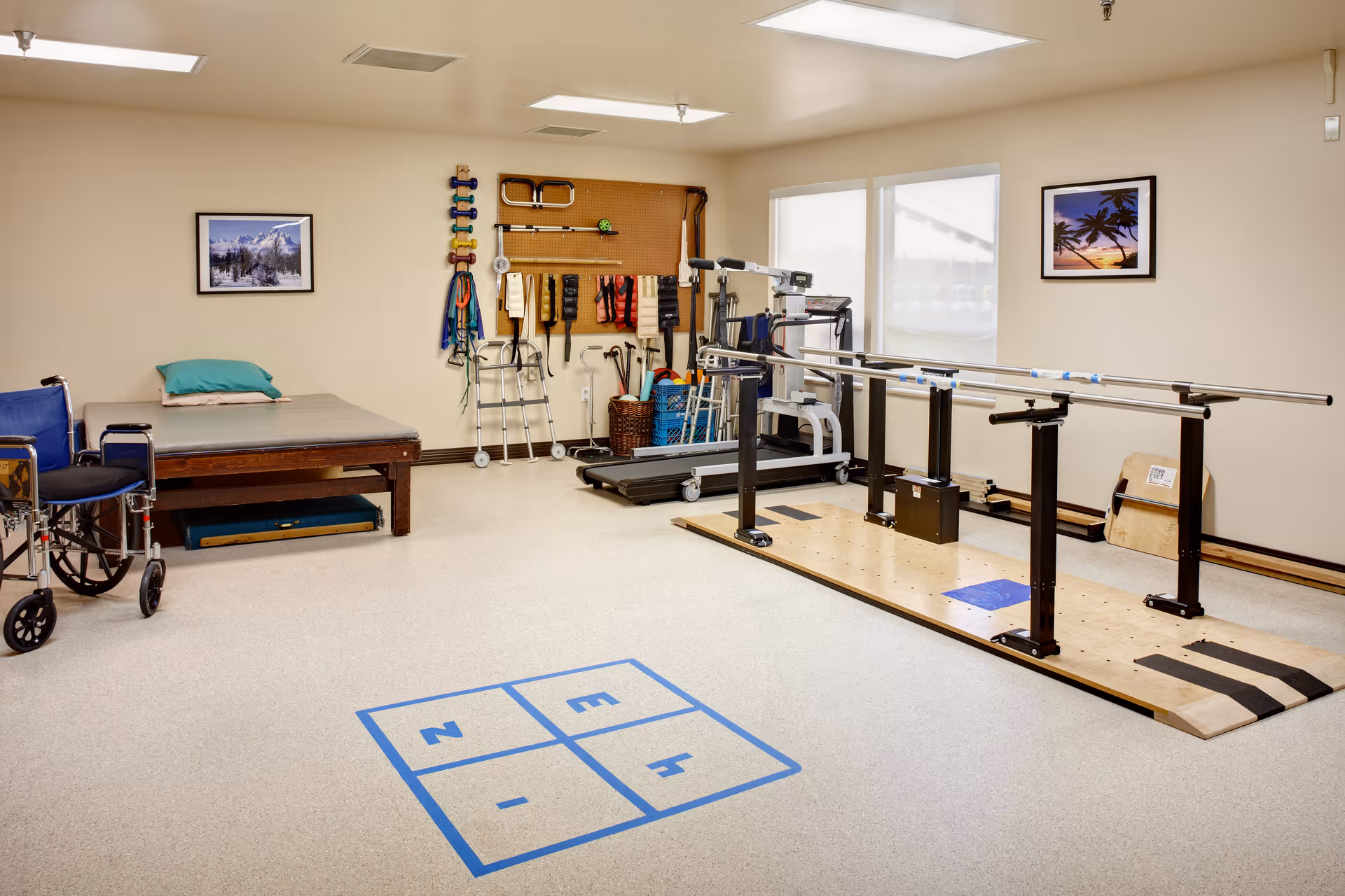 A physical therapy room with parallel bars for walking exercises, a treadmill, a wheelchair, a therapy bed with a pillow, and various exercise and therapy equipment hanging on a pegboard on the wall. The room has beige walls, two framed pictures, and a hopscotch pattern taped on the floor.