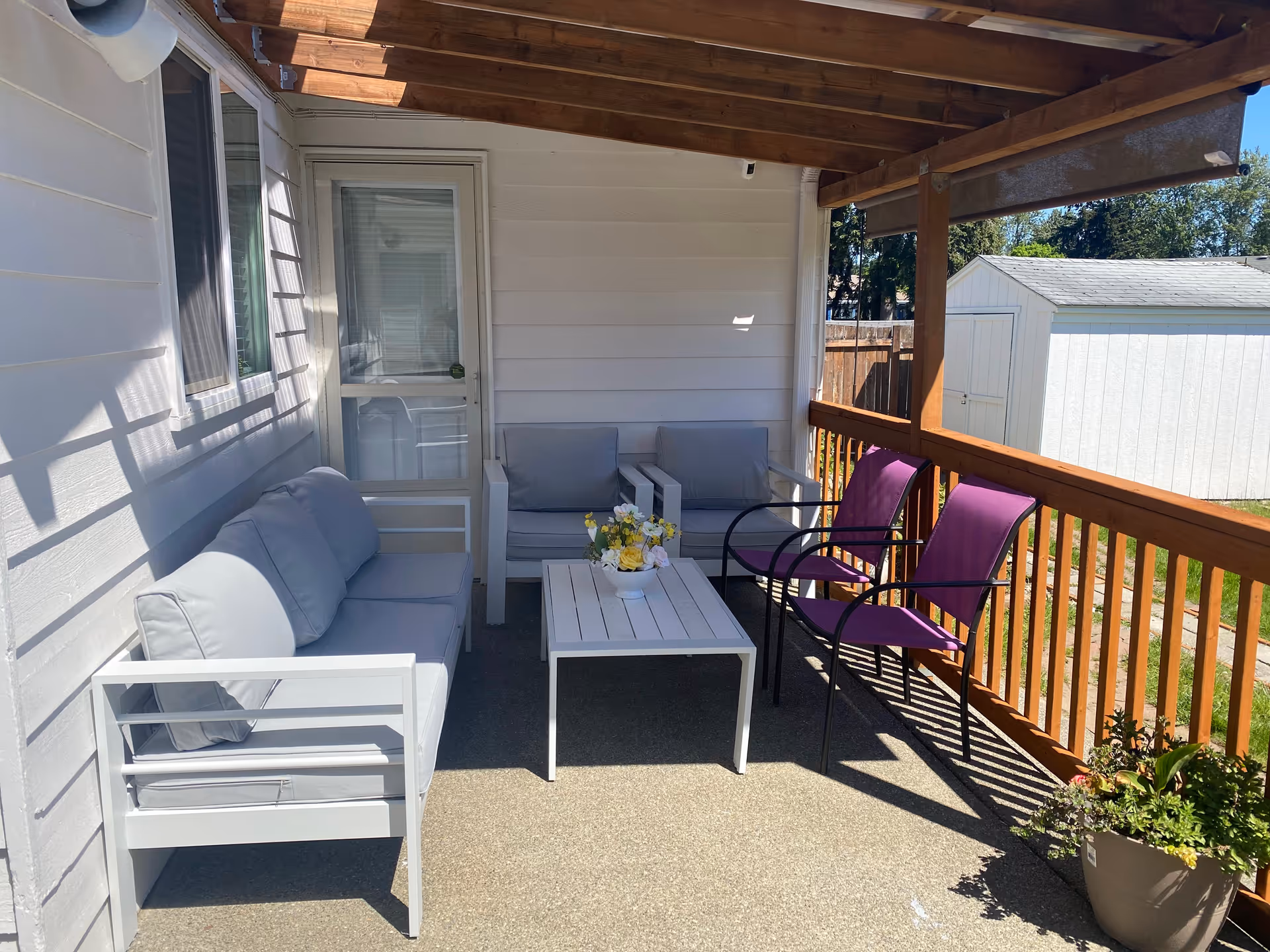 A covered outdoor patio area with a white cushioned sofa, two matching cushioned armchairs, a white coffee table with a flower arrangement, and two purple chairs. The patio has wooden railings and a wooden roof structure. A white shed and green lawn are visible in the background.