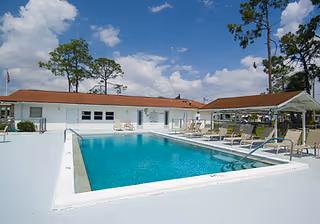 Outdoor swimming pool area with clear blue water, surrounded by white concrete deck. Several lounge chairs and tables with umbrellas are arranged around the pool. A single-story building with a red roof is visible in the background under a partly cloudy sky.