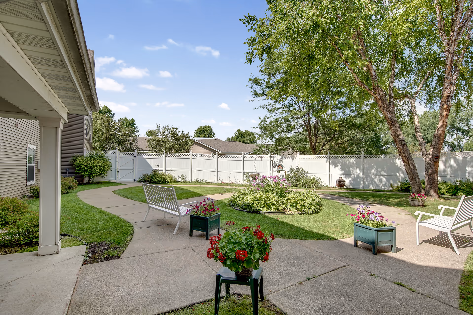 Outdoor garden area at Oak Ridge Assisted Living with a concrete pathway winding through green grass, flower beds, and trees. White benches and planters with colorful flowers are placed along the path. A white privacy fence encloses the area under a blue sky with scattered clouds.