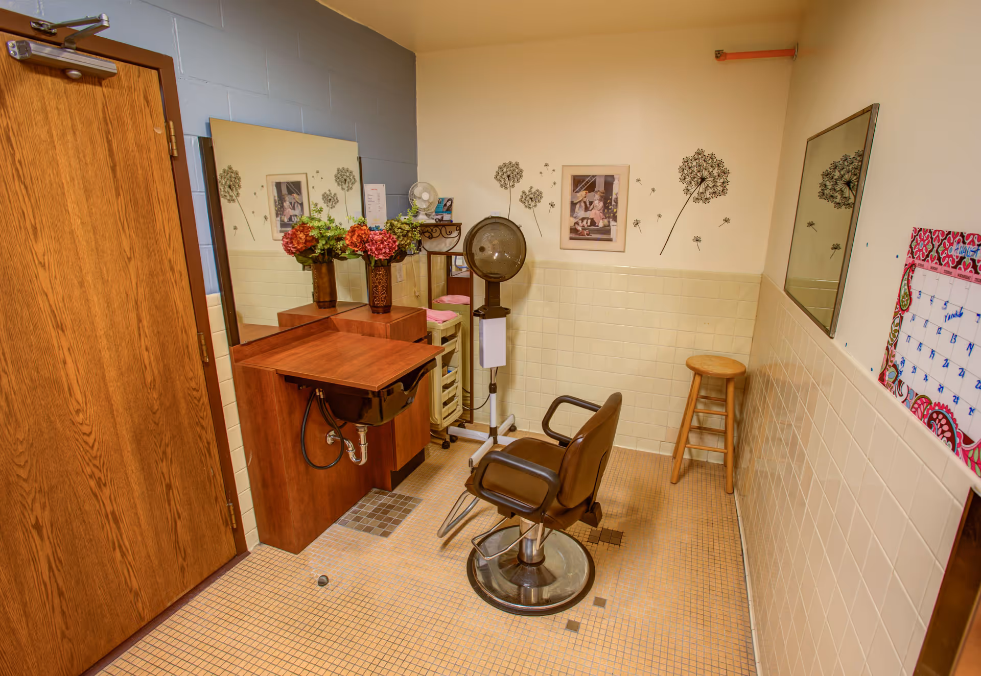 Small salon room with a styling chair, overhead hair dryer, sink, mirrors and a stool against tiled walls.