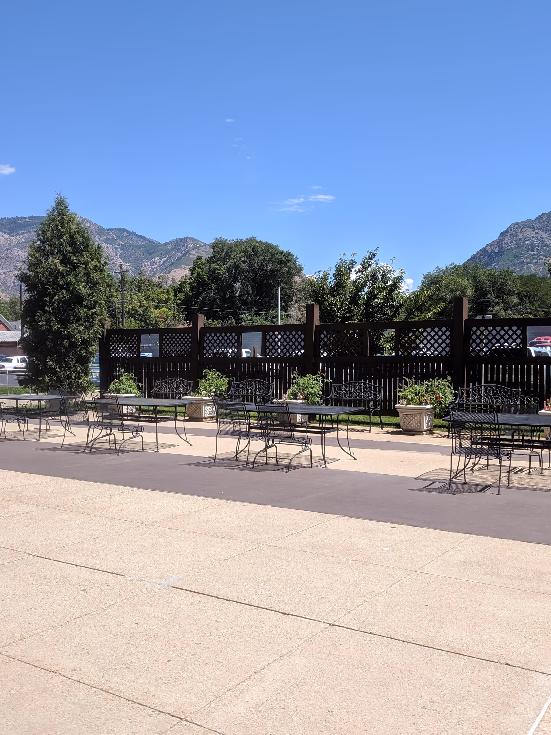 Outdoor patio area with several black metal tables and chairs arranged on a paved surface. There are decorative planters with flowers and greenery along a wooden lattice fence. In the background, there are trees, parked cars, and mountains under a clear blue sky.